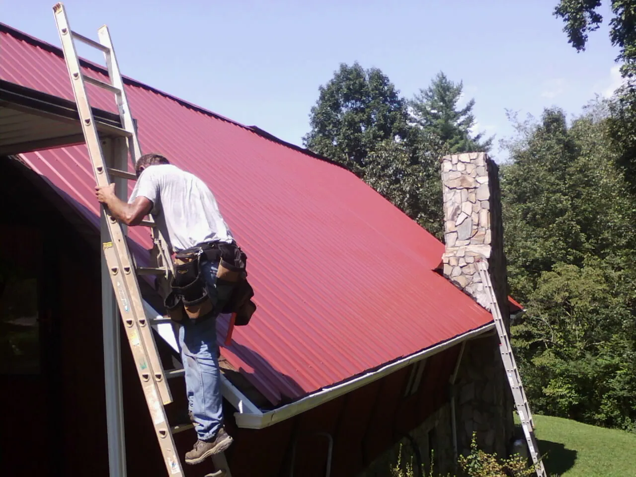 Roofer on a ladder beside a red metal roof and stone chimney — Soffit & Fascia Repair by Roofing Brothers in Charles Town